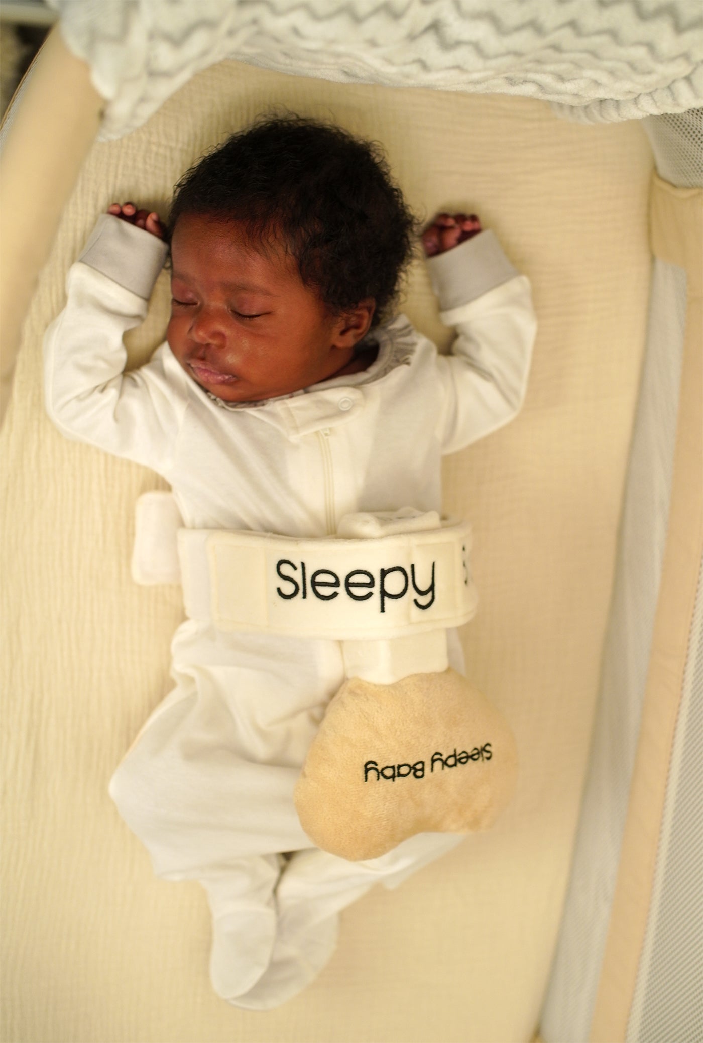 Baby in a onesie with Sleepy Baby sleep aid device attached, sleeping on a beige surface.